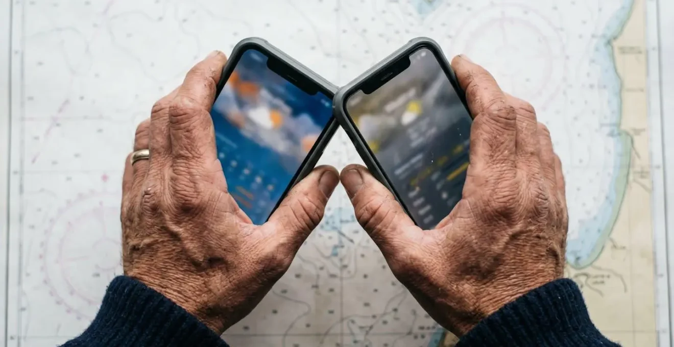Mains de navigateur tenant deux smartphones montrant des interfaces météo marine différentes, sur fond de carte nautique floue